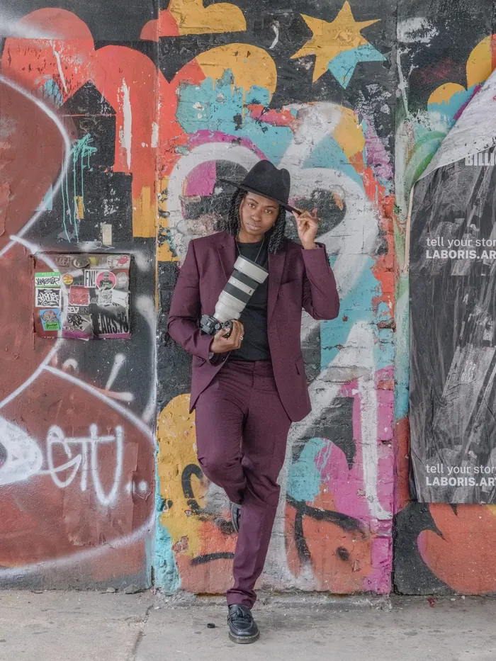 Full-length portrait of Kash in a plum suit, one hand on her hat, against a wall of SoHo graffiti