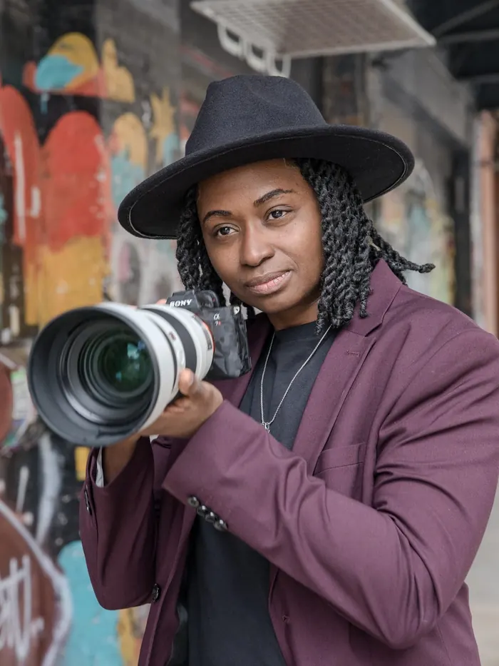 Kash in a plum suit and black hat aiming a telephoto lens toward the viewer in front of a SoHo graffiti wall